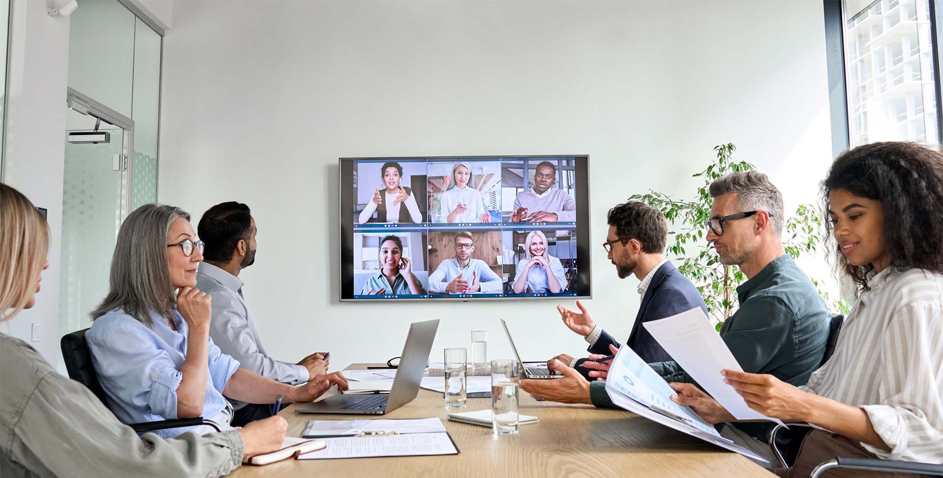 Gruppe von Menschen in einem Büro die über Barco an einer virtuellen Besprechung teilnehmen. Sechs Personen werden am Bildschirm angezeigt.